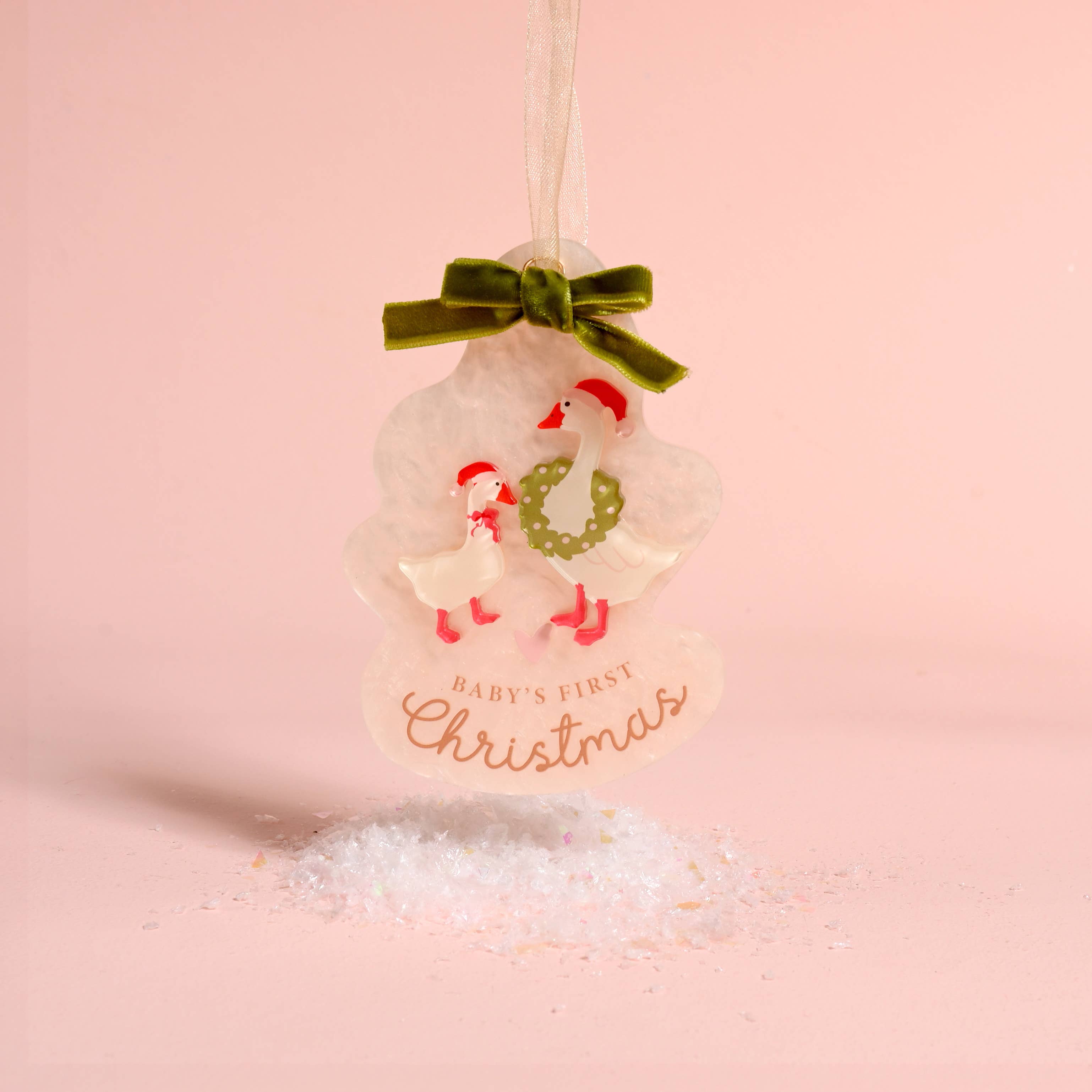 A personalized Christmas ornament shaped like a snowman with two baby swans, a green velvet bow, and the text 'BABY'S FIRST Christmas' on a soft pink background.