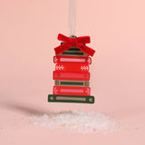 A Christmas ornament shaped like a stack of red and green books tied with a red velvet bow, hanging from a white ribbon against a soft pink background with white salt crystals at the bottom.