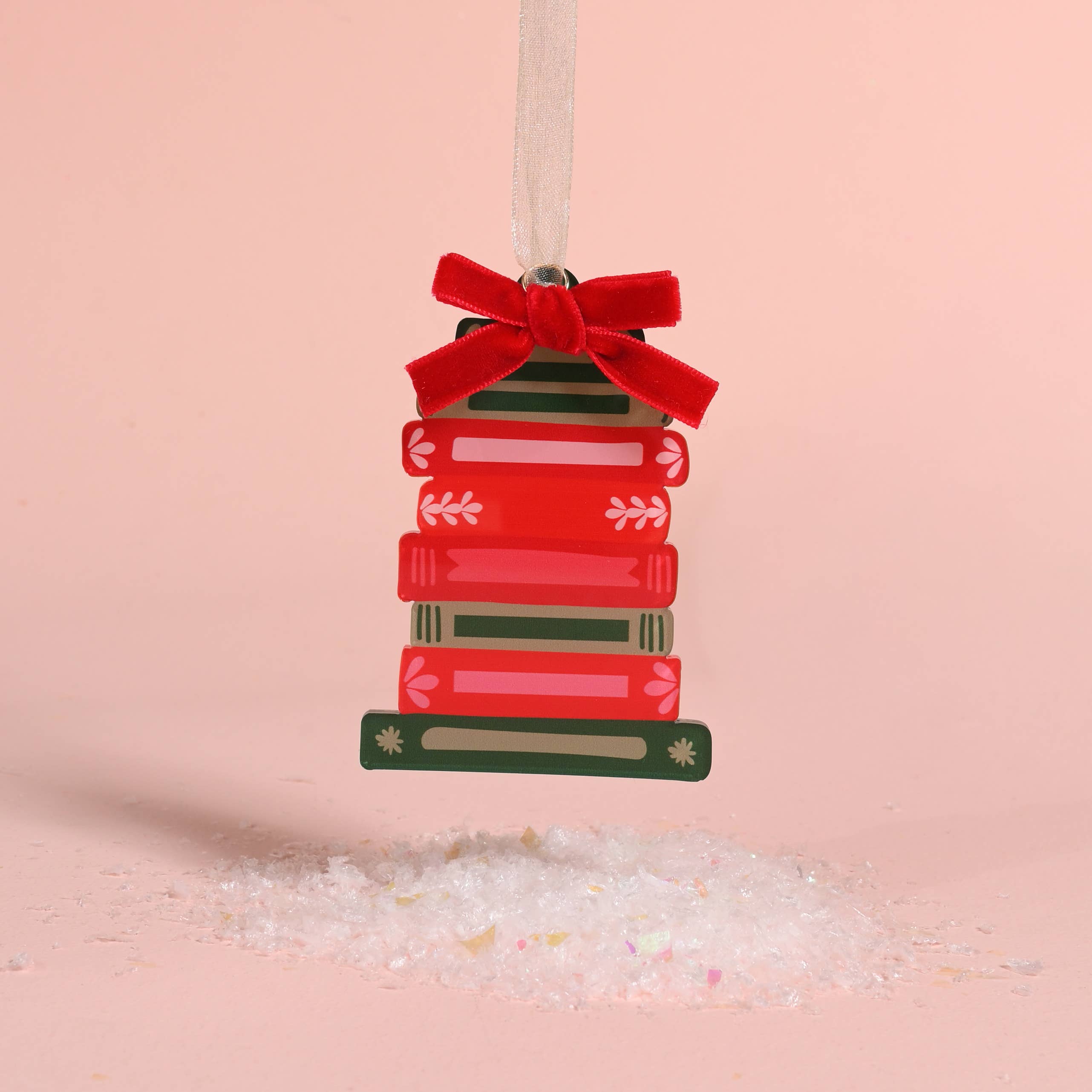 A Christmas ornament shaped like a stack of red and green books tied with a red velvet bow, hanging from a white ribbon against a soft pink background with white salt crystals at the bottom.