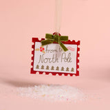 A square white holiday ornament or tag with scalloped red borders and the words 'from the North Pole' hangs from a green velvet bow. The tag is suspended over a pile of glittery white bath salts on a soft pink background.