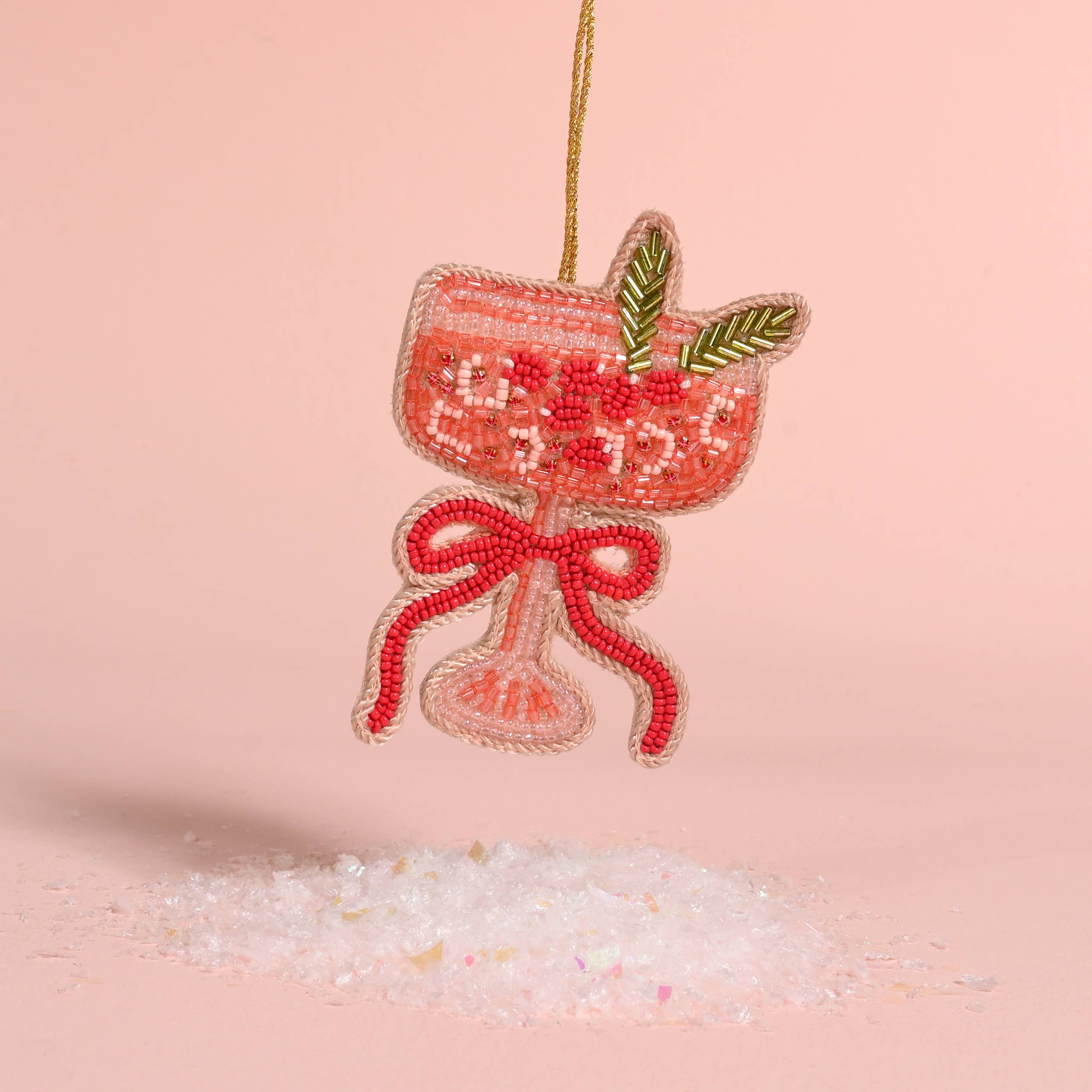 A beaded cocktail glass ornament in pink and red with green leaf accents, hanging from a gold string against a soft pink background with decorative white crystals below.