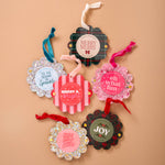 Six festive Christmas ornament tags arranged on a warm beige background. The tags feature various holiday patterns including plaid, stripes, glitter, and floral borders, with colorful ribbons and text such as 'Merry Merry', 'Merry & Bright', 'Oh what fun!', 'Joy', and 'It's the season to sparkle'.