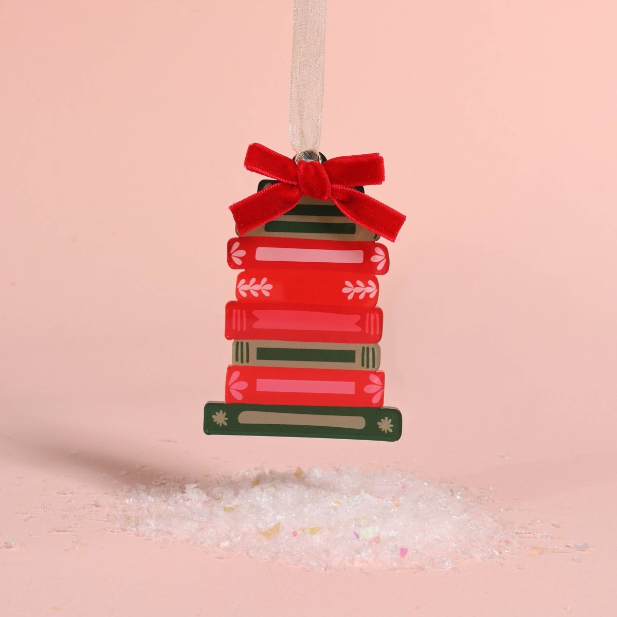 A Christmas ornament shaped like a stack of red and green books tied with a red velvet bow, hanging from a white ribbon against a soft pink background with white salt crystals at the bottom.
