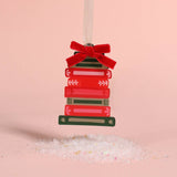 A Christmas ornament shaped like a stack of red and green books tied with a red velvet bow, hanging from a white ribbon against a soft pink background with white salt crystals at the bottom.