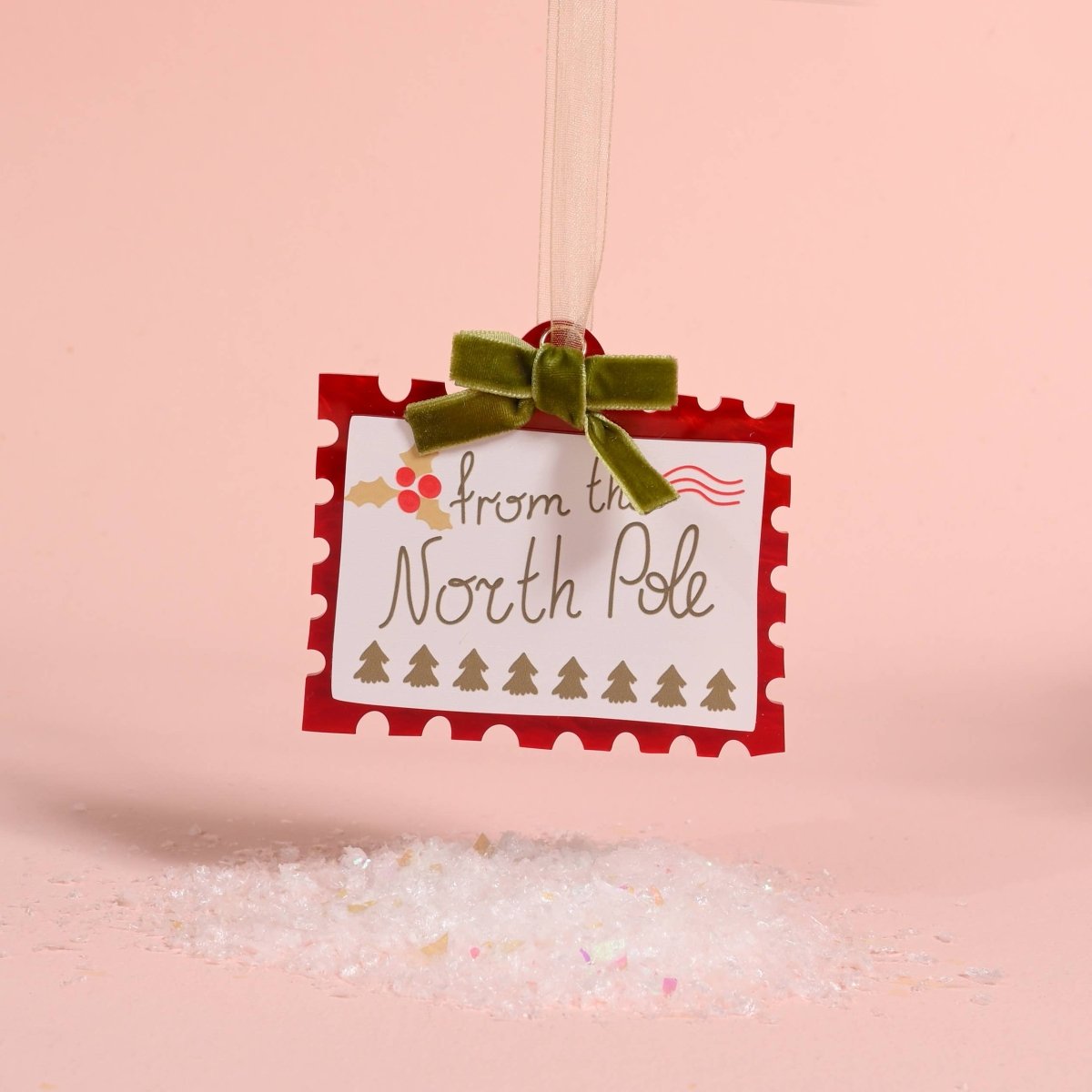 A square white holiday ornament or tag with scalloped red borders and the words 'from the North Pole' hangs from a green velvet bow. The tag is suspended over a pile of glittery white bath salts on a soft pink background.