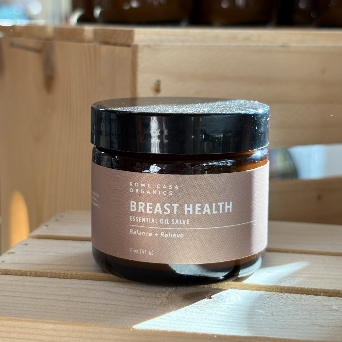 An amber glass jar of Rowe Casa Organics Breast Health Essential Oil Salve with a black lid, labeled “Balance + Relieve, 2 oz,” resting on a light wooden surface with warm natural light and softly blurred wooden boxes in the background.