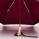 Maroon duckhead umbrella with wooden handle and strap on a light pink background