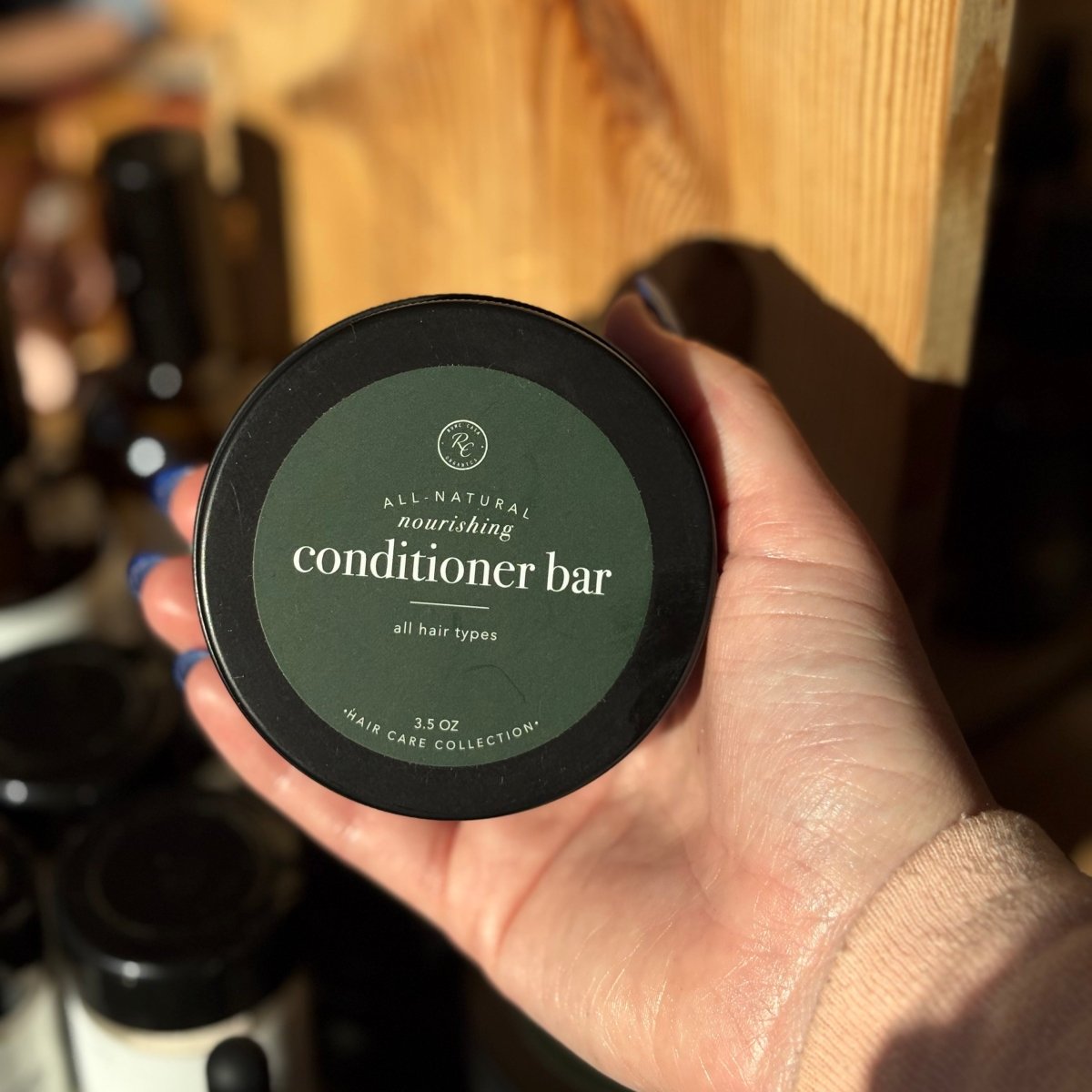 Hand holding a container of All-Natural Nourishing Conditioner Bar with a wooden shelf in the background.