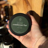 Hand holding a container of All-Natural Nourishing Conditioner Bar with a wooden shelf in the background.