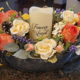 Decorative floral arrangement with a candle labeled 'Forever Loved' on a dark surface.