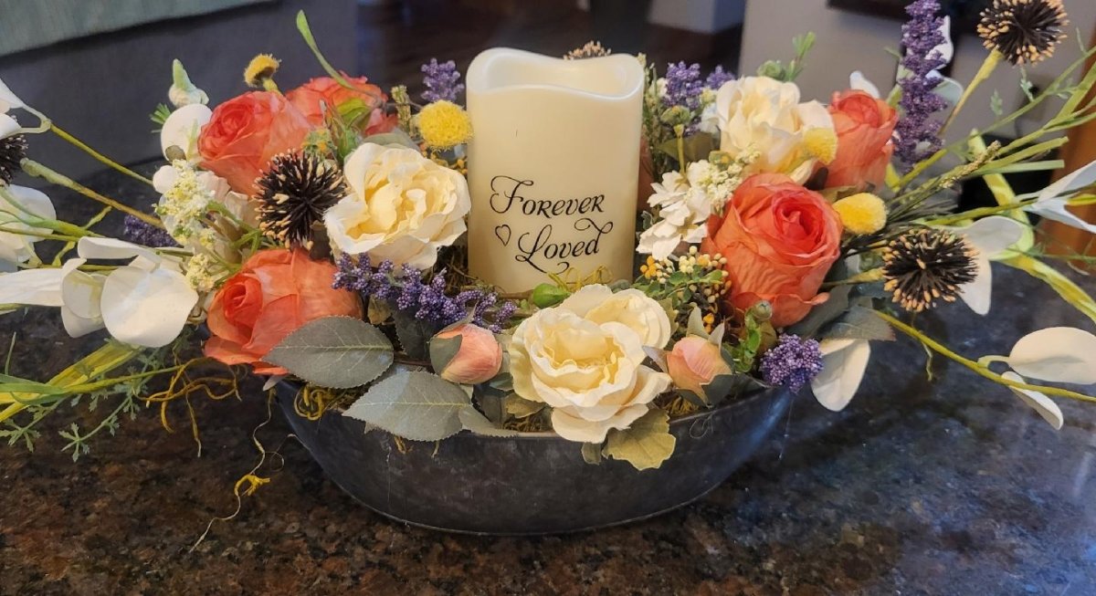 Decorative floral arrangement with a candle labeled 'Forever Loved' on a dark surface.