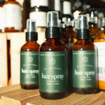 Three bottles of All Natural Hair Care Collection hair spray on a wooden surface with shelves in the background.
