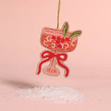 A beaded cocktail glass ornament in pink and red with green leaf accents, hanging from a gold string against a soft pink background with decorative white crystals below.
