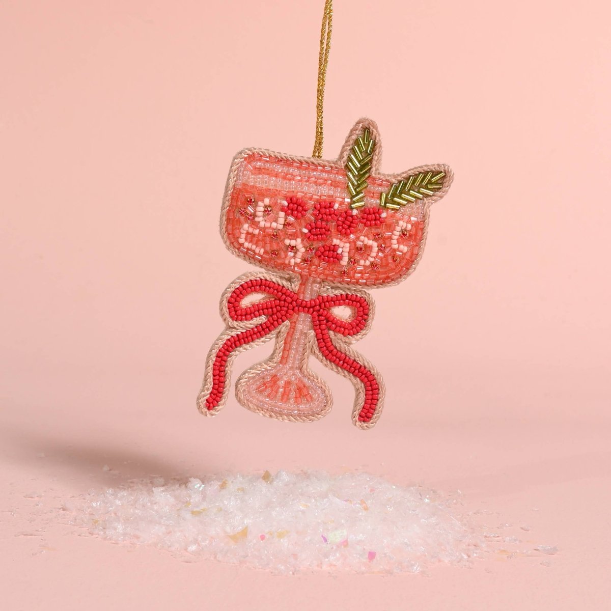 A beaded cocktail glass ornament in pink and red with green leaf accents, hanging from a gold string against a soft pink background with decorative white crystals below.