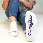 White socks with 'courageous' text worn by a person sitting on a white floor.