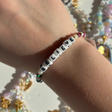 Little Words Project Bracelet with 'BELIEVE' beads on a wrist with glittery background
