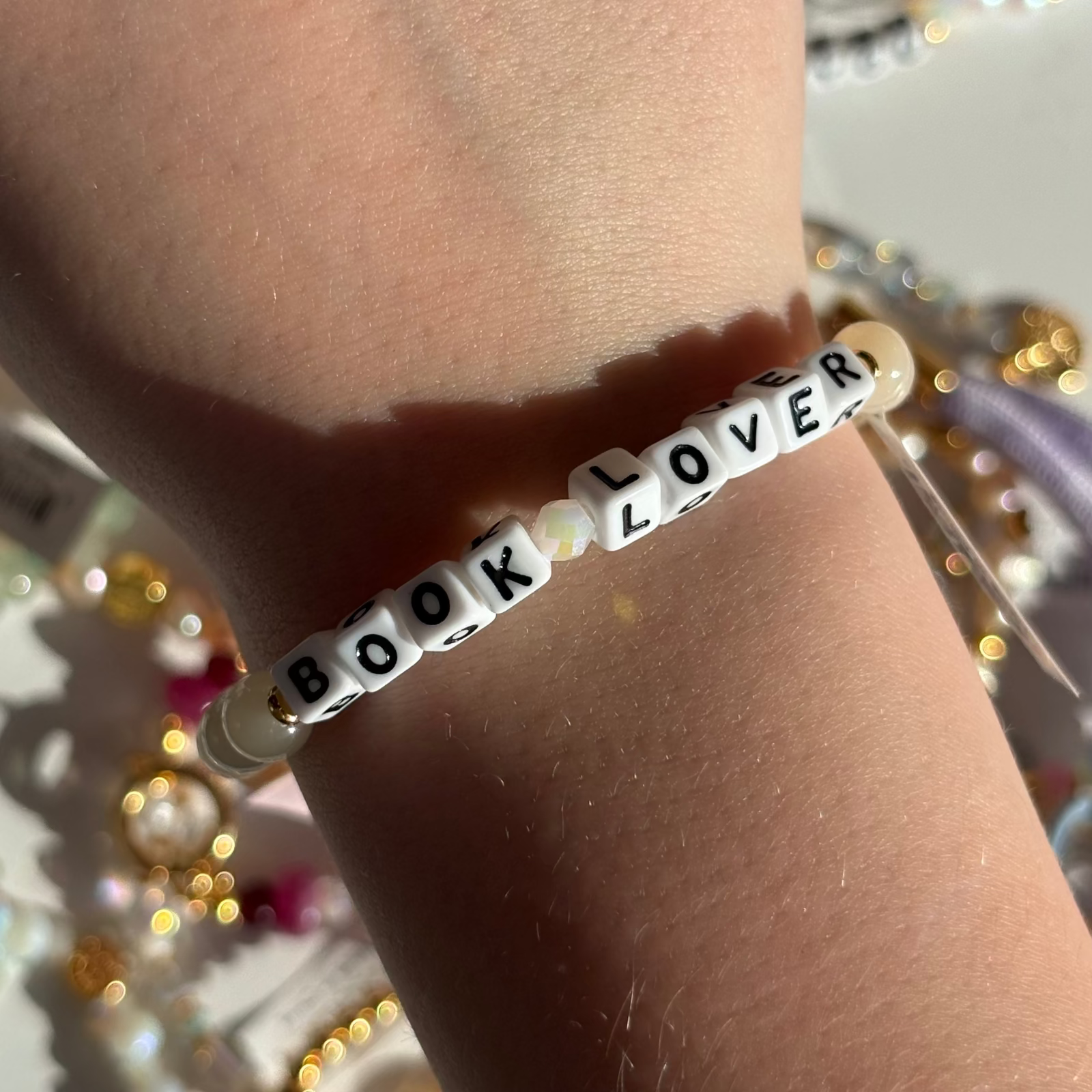 Little Words Project Bracelet with 'BOOK LOVER' beads on a wrist against a glittery background