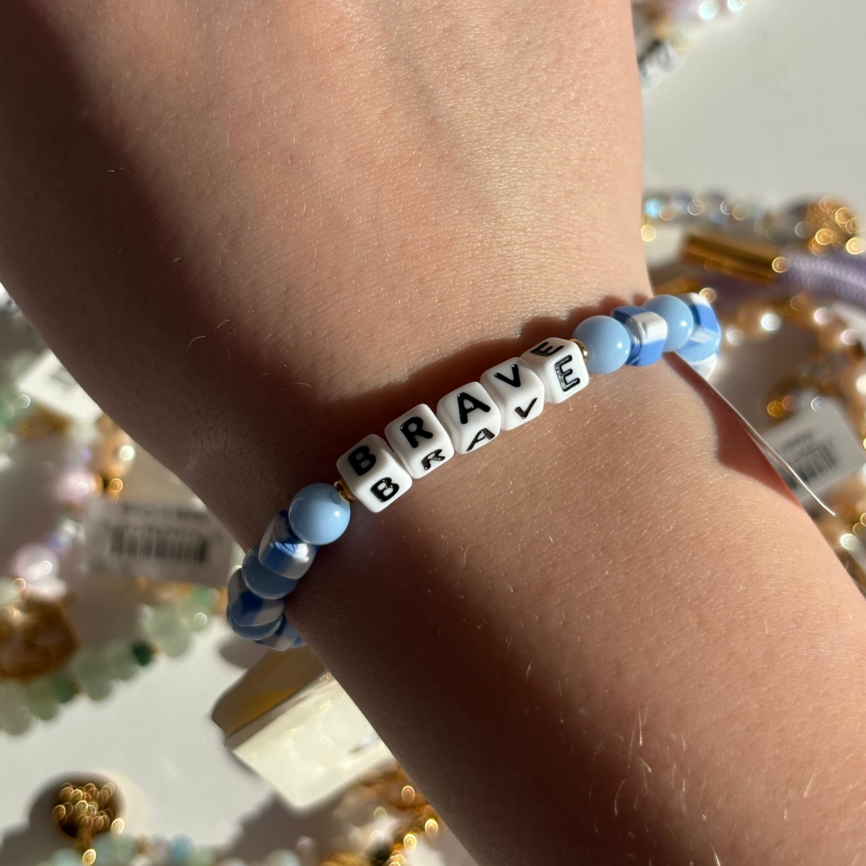 Little Words Project Bracelet with 'BRAVE' beads on a wrist, with a blurred background of colorful beads.