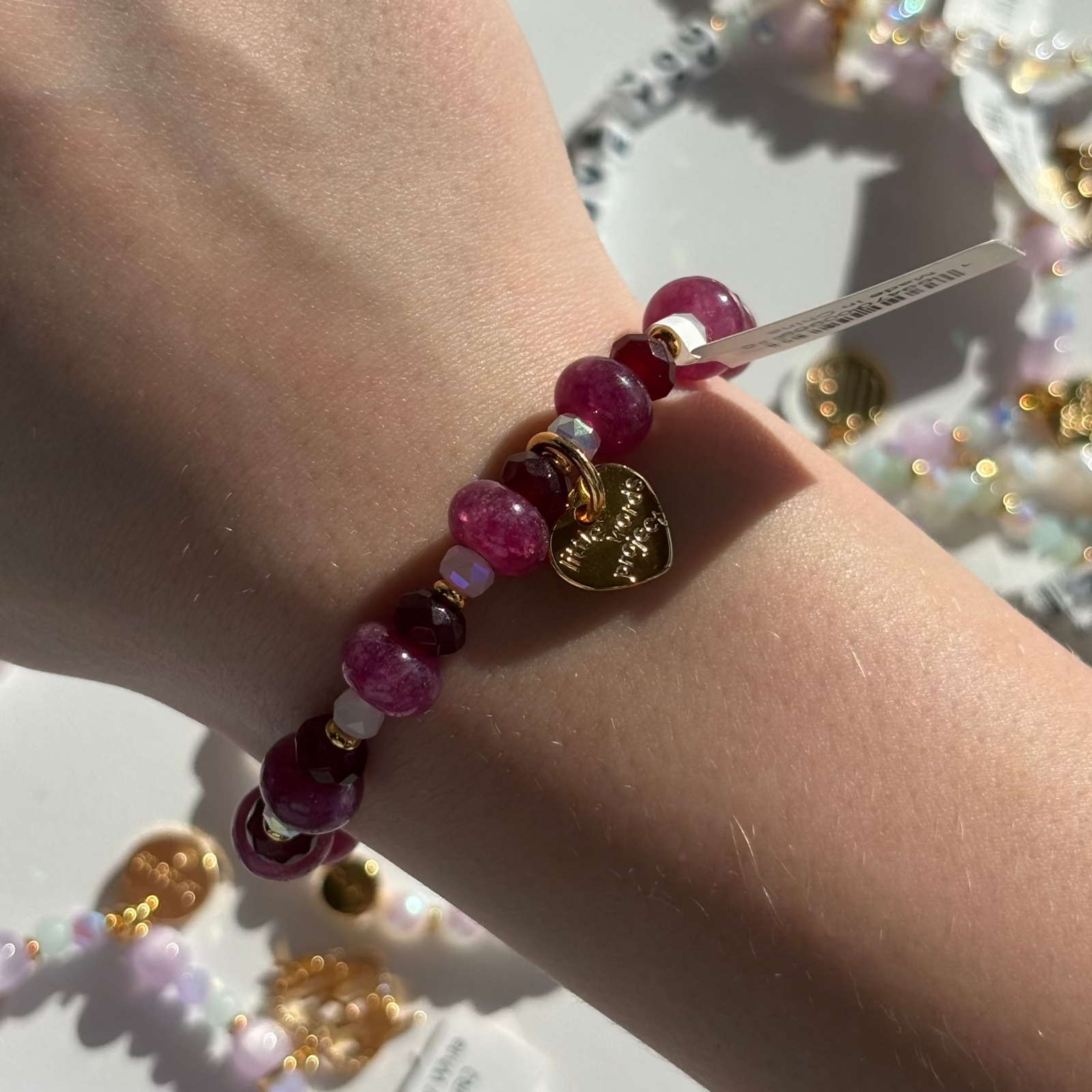 Little Words Project Bracelet with red beads and gold accents on a wrist, with a blurred background of more jewelry.