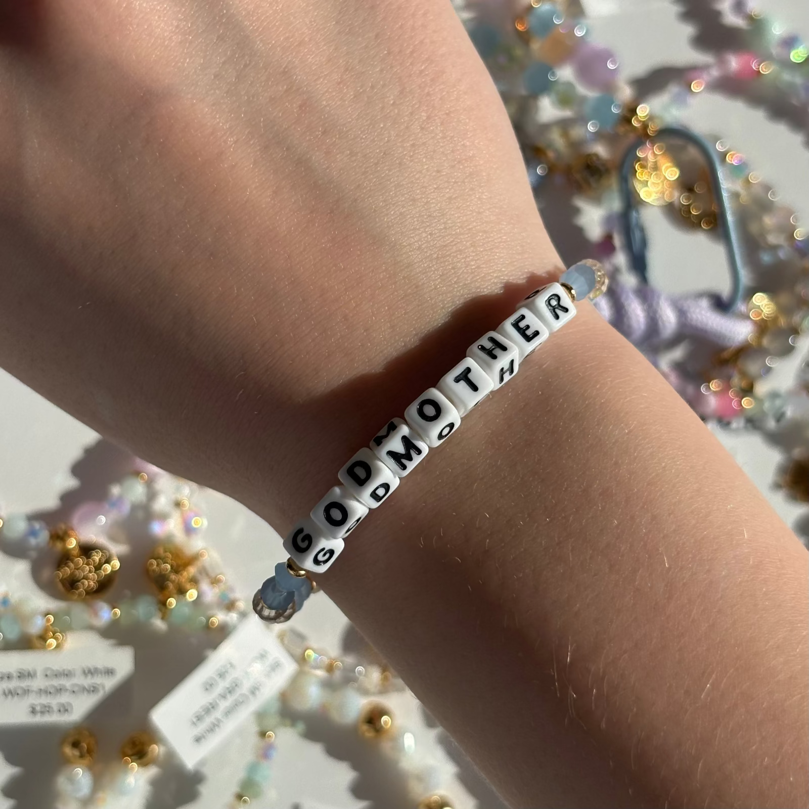 Little Words Project Bracelet with 'GODMOTHER' beads on a wrist, surrounded by decorative items.
