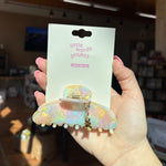Hand holding a colorful hair clip teddy bear charm with 'little words project' packaging.