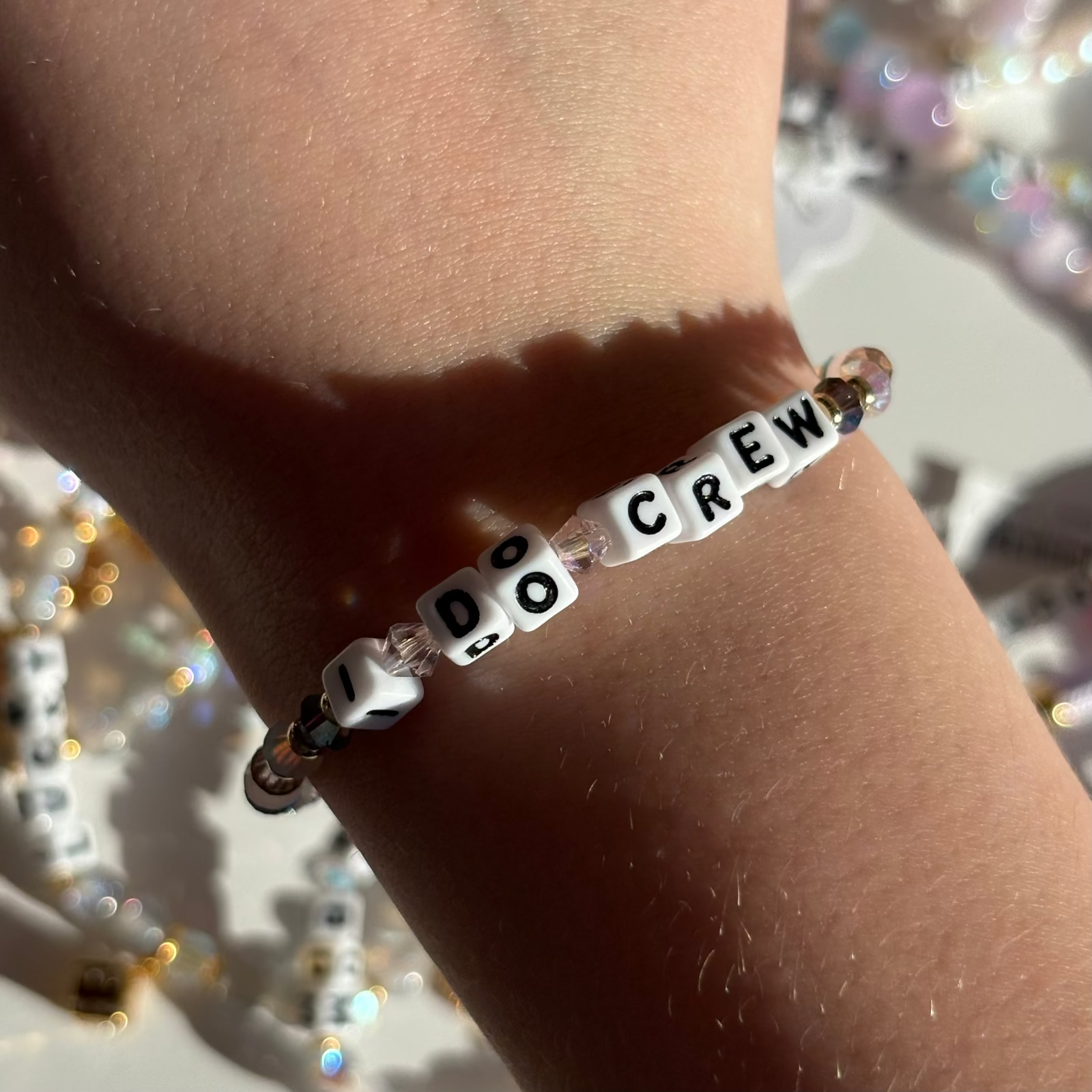Little Words Project Bracelet with 'I DO CREW' beads on a wrist, with a blurred background of colorful beads.