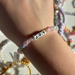 Little Words Project Bracelet with 'JOY' beads on a wrist, with a blurred background of colorful beads.