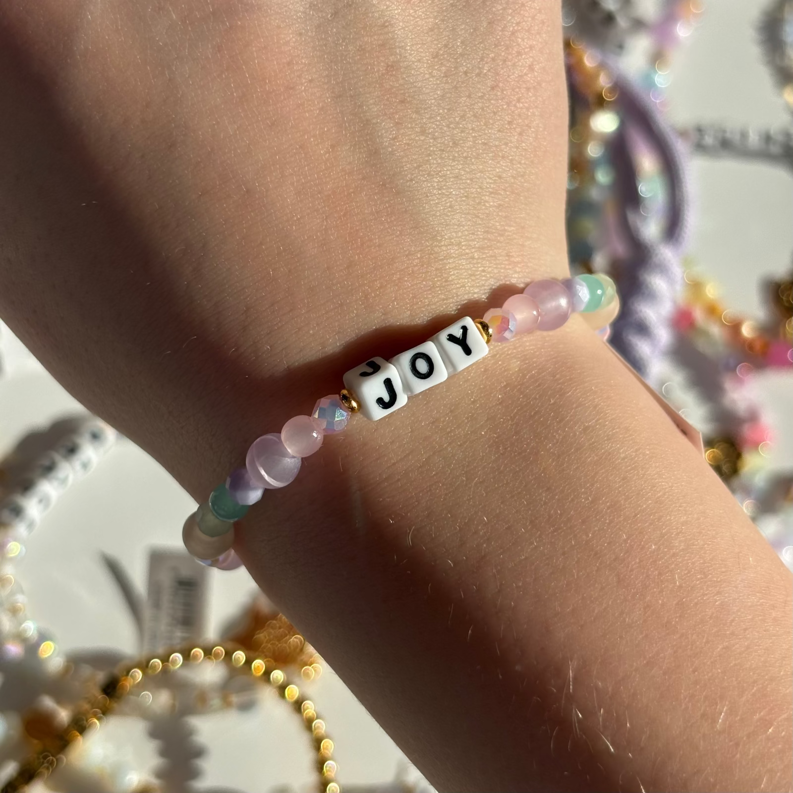 Little Words Project Bracelet with 'JOY' beads on a wrist, with a blurred background of colorful beads.