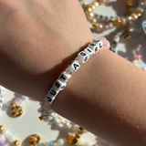 Little Words Project Bracelet with 'JUST A GIRL' beads on a wrist, with a blurred decorative background.