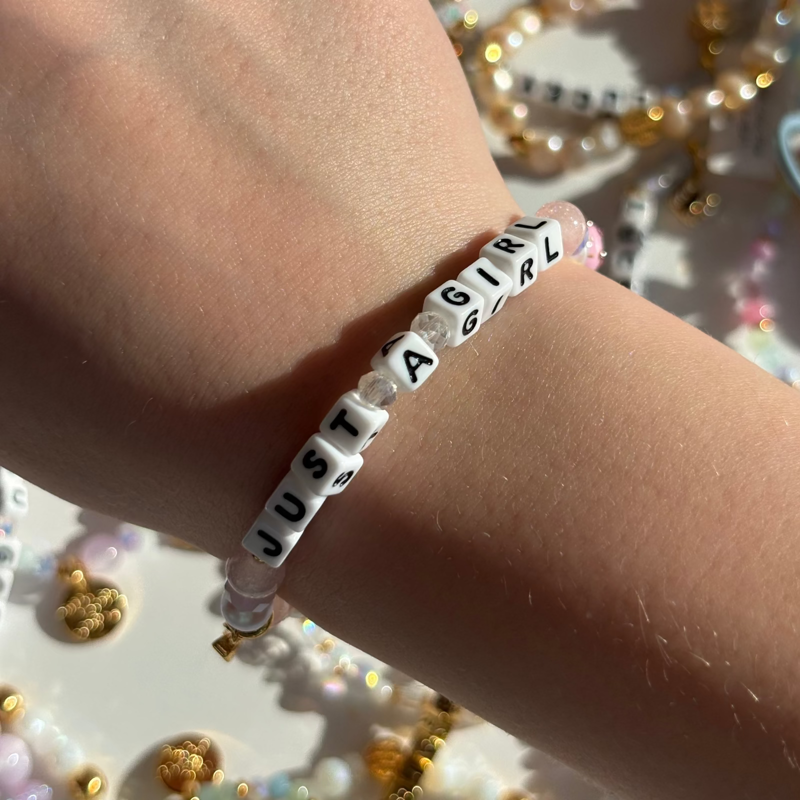 Little Words Project Bracelet with 'JUST A GIRL' beads on a wrist, with a blurred decorative background.