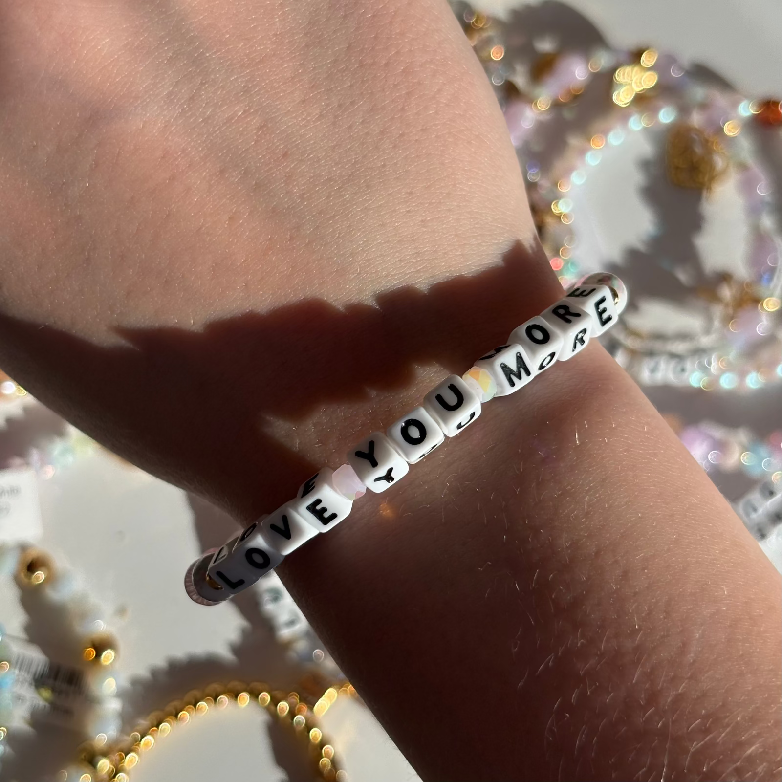 Little Words Project Bracelet with 'LOVE YOU MORE' on a wrist against a glittery background