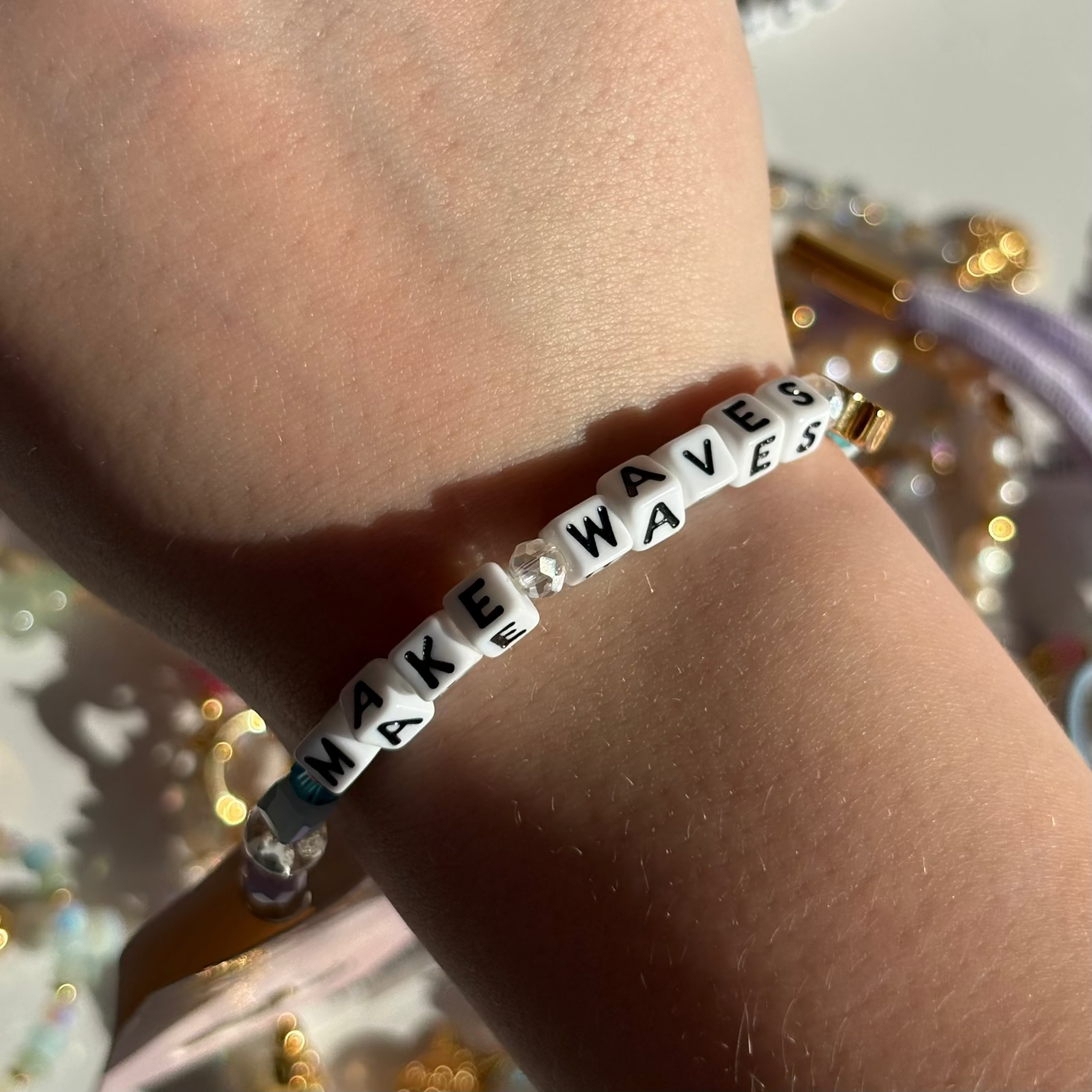 Little Words Project Bracelet with 'MAKE WAVES' beads on a wrist against a glittery background