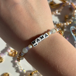Little Words Project Bracelet with 'MAMA' beads on a wrist, surrounded by gold jewelry on a light background