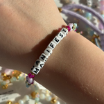 Little Words Project Bracelet with 'MANIFEST' beads on a wrist, surrounded by colorful beads.