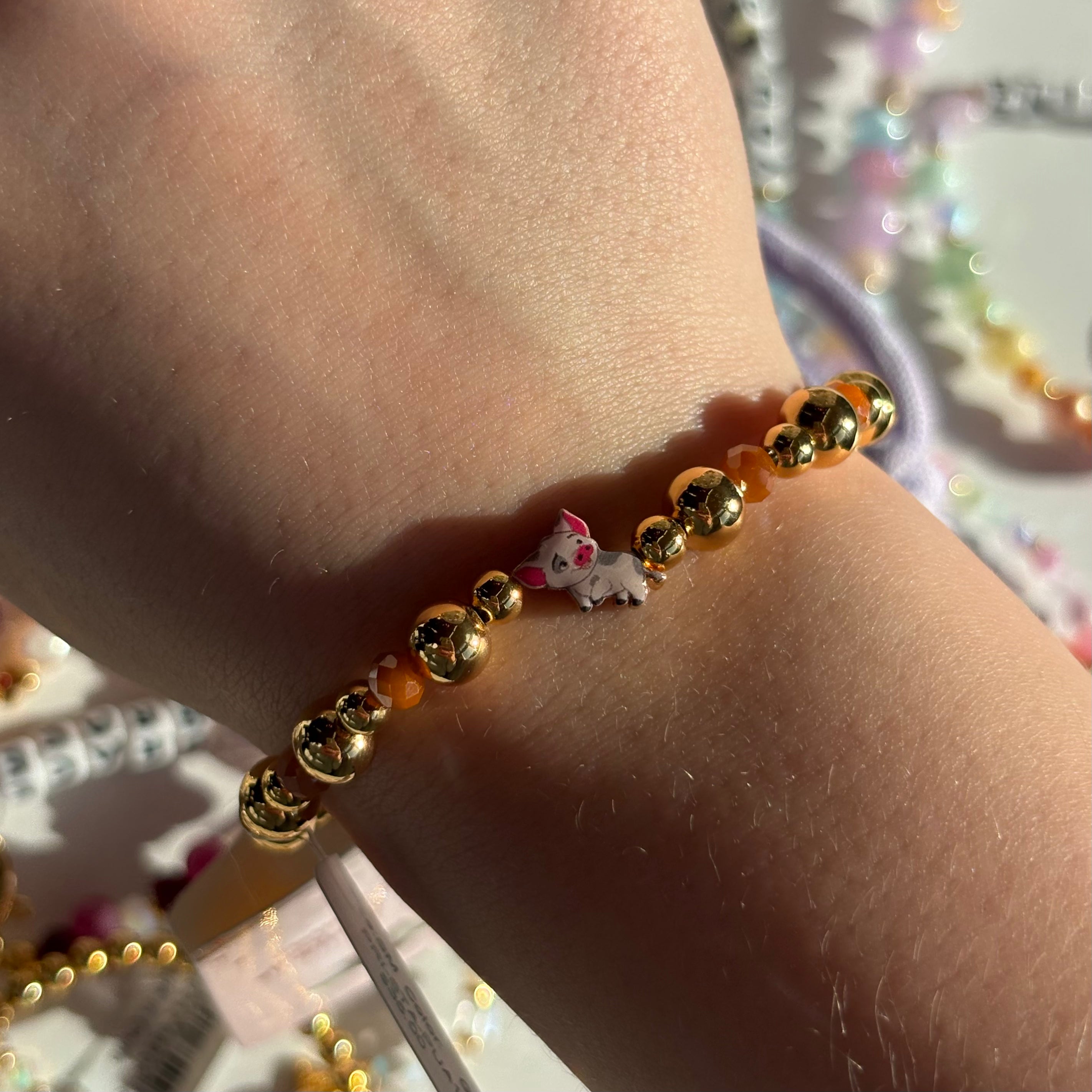 Gold beaded bracelet with a small pig named pua from moana disney charm on a wrist, with a blurred colorful background.