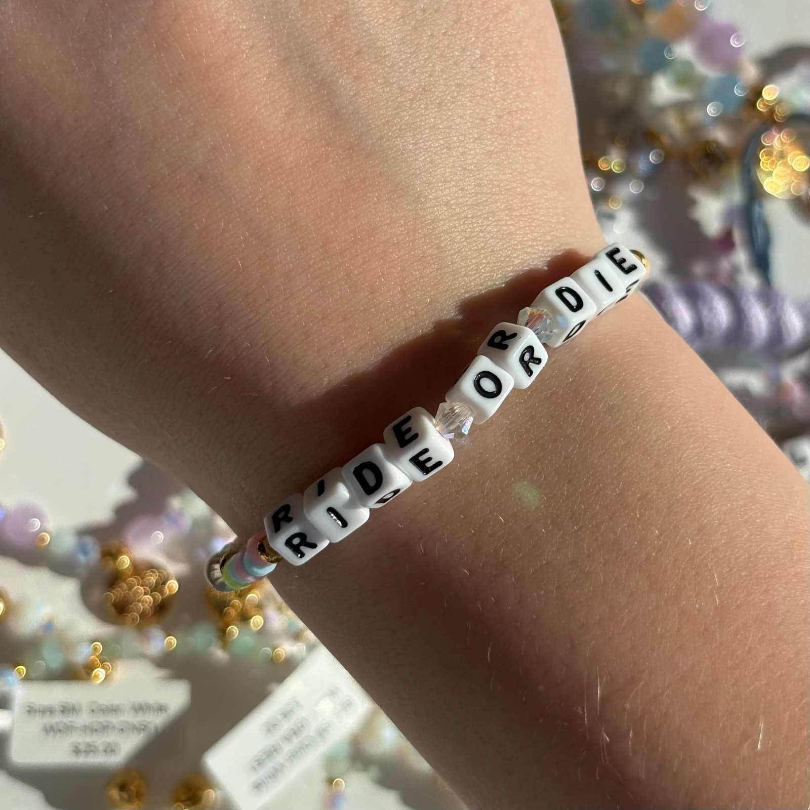 Little Words Project Bracelet with 'RIDE OR DIE' beads on a wrist, with a blurred background of colorful beads.