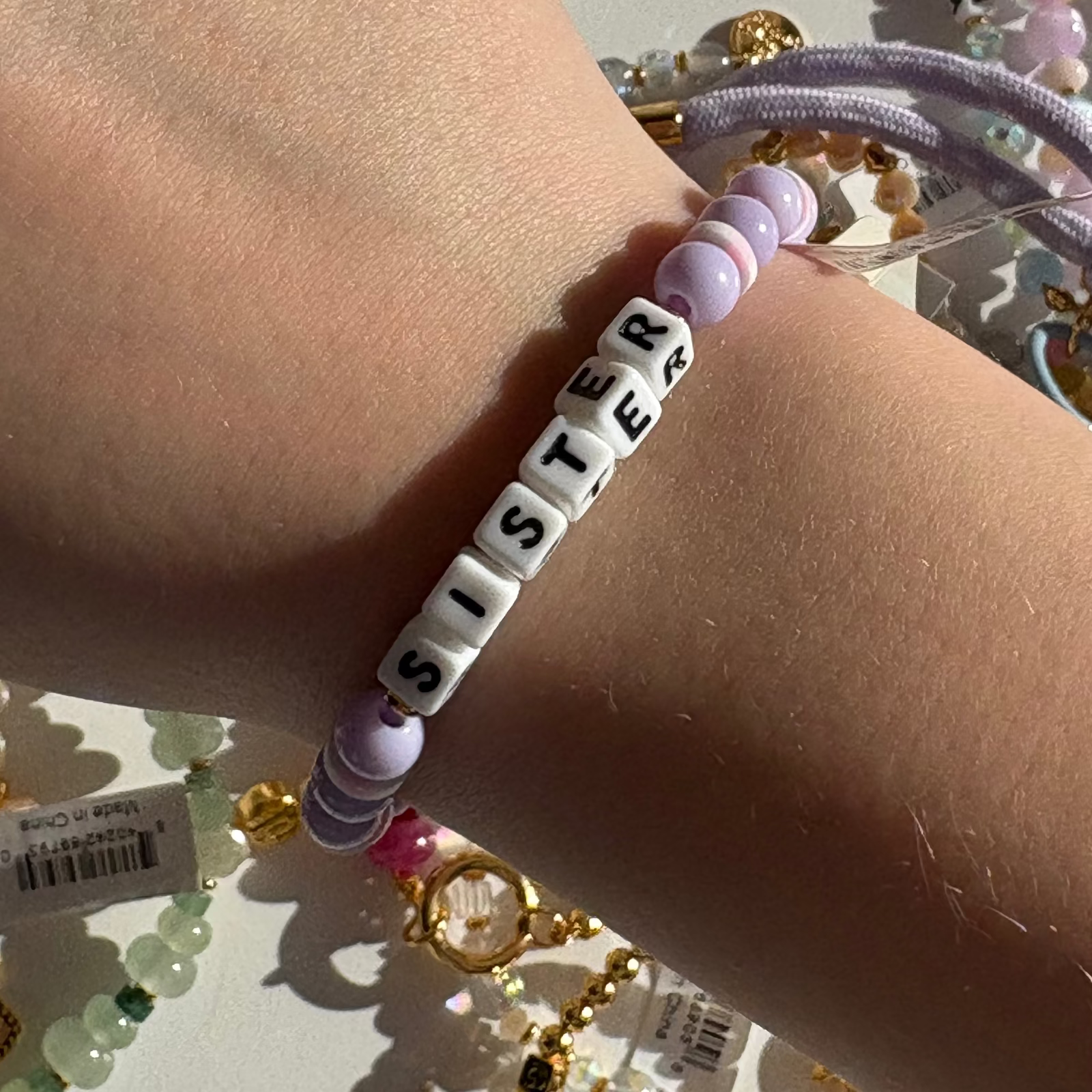 Little Words Project Bracelet with 'SISTER' beads on a wrist, with jewelry in the background
