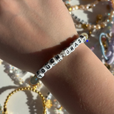 Little Words Project Bracelet with 'TOUCH GRASS' on a wrist with a blurred background