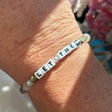Bracelet with 'LET THEM' beads on a wrist with a blurred background