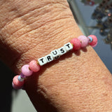 LWP bracelet with pink beads and a white tile spelling 'TRUST' on a wrist.