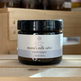 A small amber glass jar of Rowe Casa Mama’s Milk Salve with a black lid, labeled “All-Natural Baby Mama’s Milk Salve – boost + supply, 2 fl oz,” displayed on a light wooden surface with softly blurred wooden boxes in the background.
