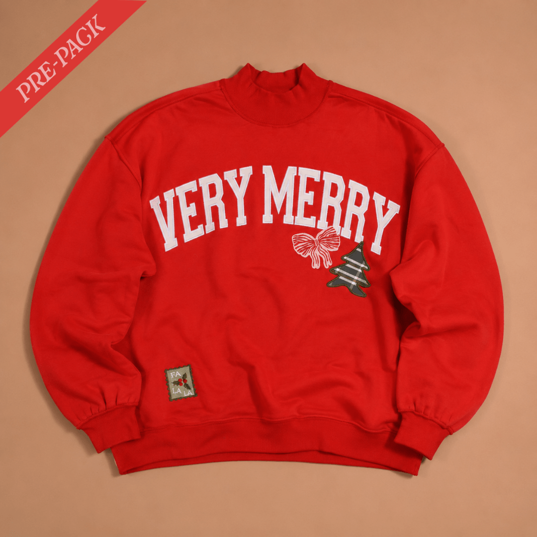 A red crewneck sweatshirt with 'VERY MERRY' printed in white arched letters across the chest, accompanied by a small bow and Christmas tree graphic. The sweatshirt is laid flat on a beige background with a red 'PRE-PACK' banner in the top-left corner.