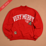 A red crewneck sweatshirt with 'VERY MERRY' printed in white arched letters across the chest, accompanied by a small bow and Christmas tree graphic. The sweatshirt is laid flat on a beige background with a red 'PRE-PACK' banner in the top-left corner.