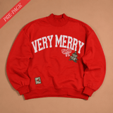 A red crewneck sweatshirt with 'VERY MERRY' printed in white arched letters across the chest, accompanied by a small bow and Christmas tree graphic. The sweatshirt is laid flat on a beige background with a red 'PRE-PACK' banner in the top-left corner.