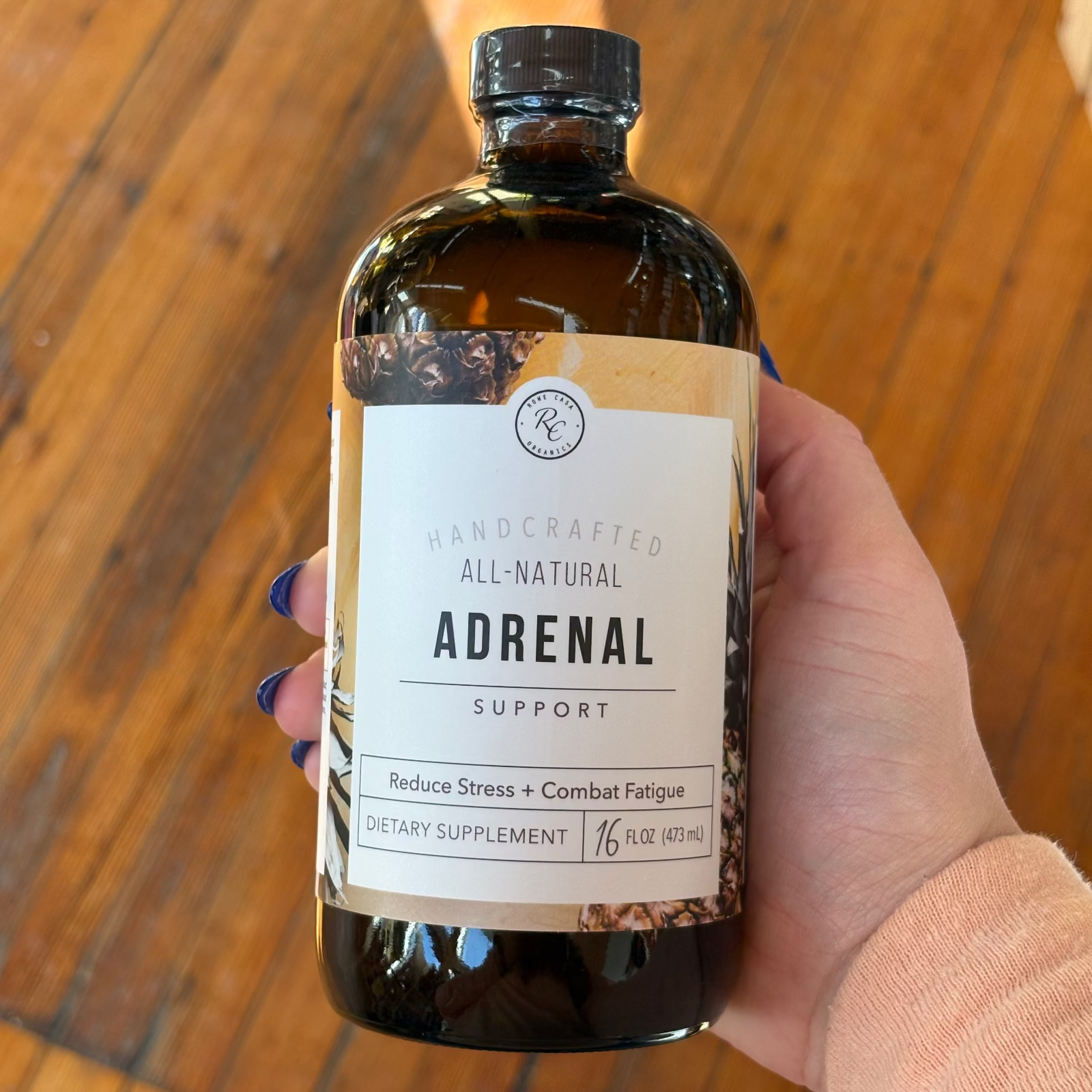 Rowe Casa hand holding a bottle labeled 'Adrenal Support' against a wooden background