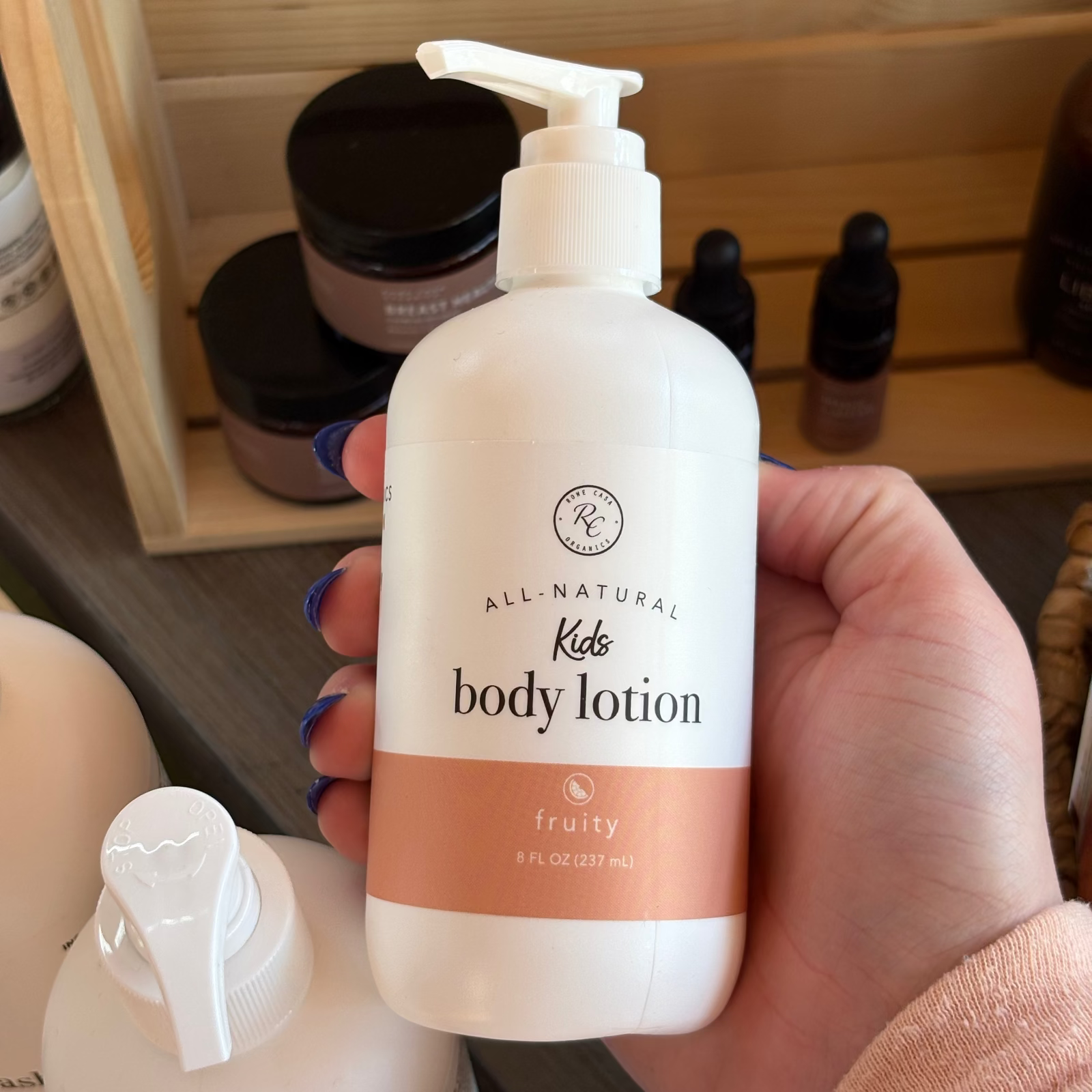 Hand holding a bottle of  Rowe Casa All-Natural Kids Body Lotion' with a wooden shelf in the background.