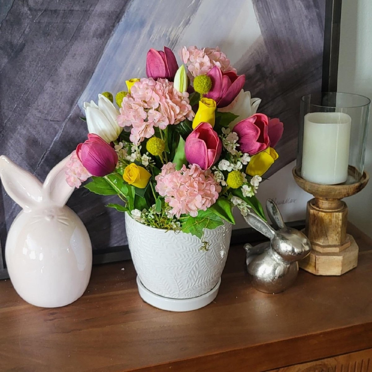Floral arrangement in a white pot on a wooden surface with decorative items.