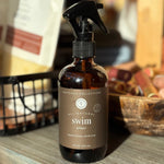 Brown spray bottle labeled 'All Natural Swim Spray' on a wooden surface with a blurred background.