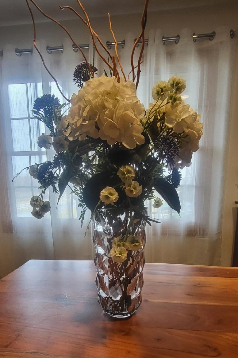 Floral arrangement in a vase on a wooden table with a window and curtain in the background