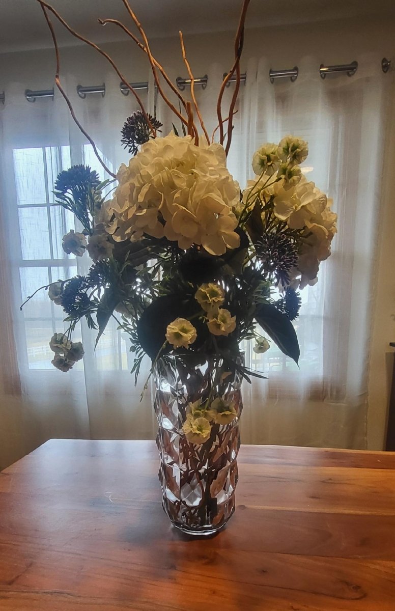 Floral arrangement in a vase on a wooden table with a window and curtain in the background